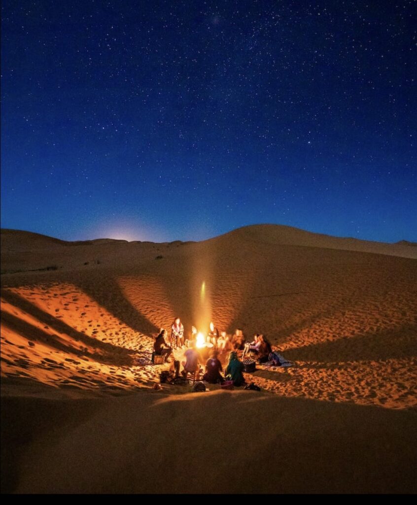 People sitting around a campfire on the beach looking at the stars in the sky. The sun is setting in the distance.